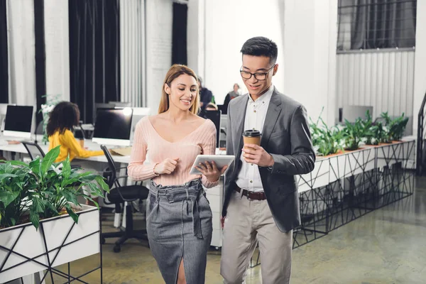 Colegas de negocios sonrientes utilizando tableta digital y caminando en la oficina - foto de stock