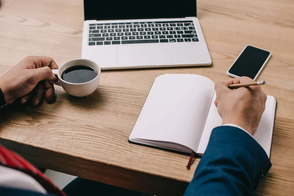 Ausgeschnittene Ansicht eines Geschäftsmannes, der am Arbeitsplatz mit Kaffeetasse und digitalen Geräten in Notizbuch schreibt — Stockfoto