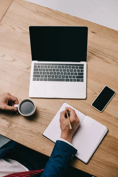 Vista recortada de hombre de negocios escribiendo en cuaderno en el lugar de trabajo con café y gadgets — Stock Photo