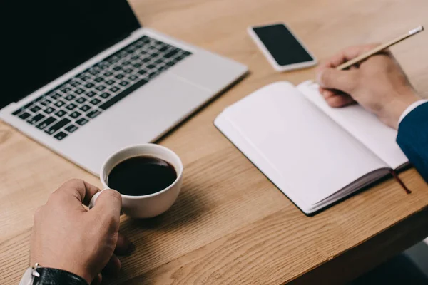 Vista recortada del hombre de negocios sosteniendo taza de café mientras escribe en el cuaderno en el lugar de trabajo - foto de stock