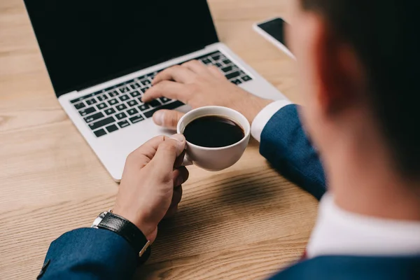 Vista recortada de hombre de negocios sosteniendo la taza de café mientras escribe en el ordenador portátil en la mesa — Stock Photo