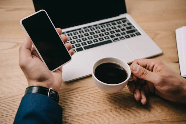 Vista recortada de empresario sosteniendo teléfono inteligente y taza de café en el lugar de trabajo con el ordenador portátil - foto de stock