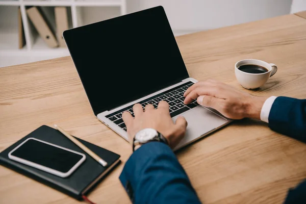 Vista cortada de empresário digitando no laptop no local de trabalho com smartphone e xícara de café — Fotografia de Stock