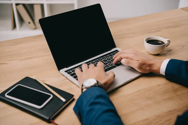 Ausgeschnittene Ansicht eines Geschäftsmannes, der am Arbeitsplatz mit Smartphone und Tasse Kaffee am Laptop tippt — Stockfoto