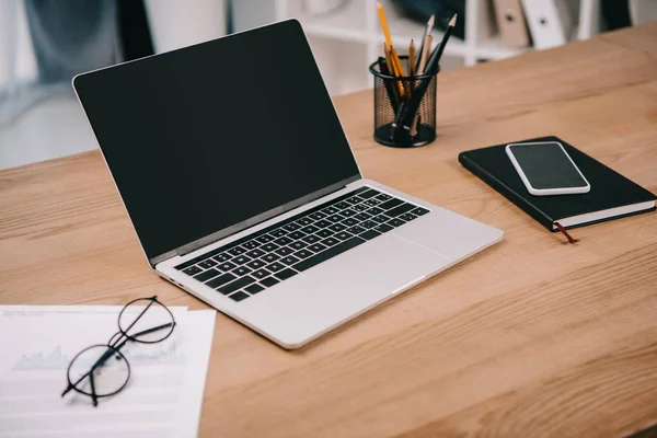 Portátil con pantalla en blanco, documentos comerciales, teléfonos inteligentes y suministros de oficina en el lugar de trabajo - foto de stock