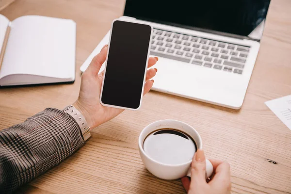 Vue recadrée de femme d'affaires en utilisant un smartphone et tenant une tasse de café sur le lieu de travail avec ordinateur portable — Photo de stock