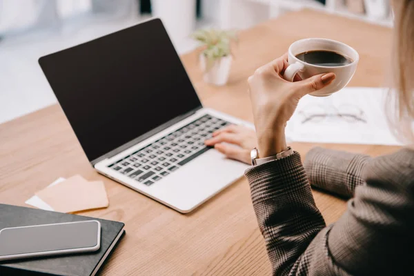 Обрезанный вид деловой женщины, держащей чашку кофе во время работы с ноутбуком в офисе — стоковое фото