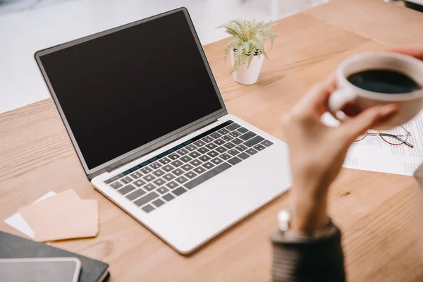 Vista recortada de la mujer de negocios sosteniendo la taza de café mientras está sentado en el lugar de trabajo con el ordenador portátil - foto de stock