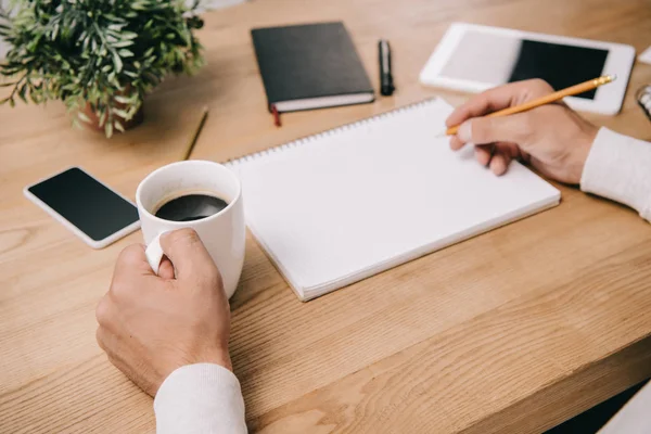 Visão cortada de empresário com xícara de café escrevendo em notebook no local de trabalho com gadgets — Fotografia de Stock