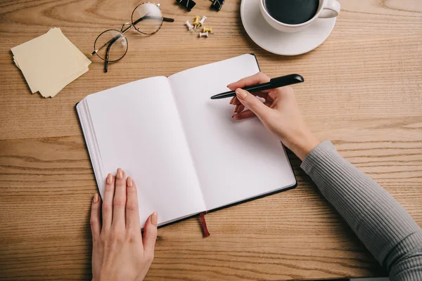 Vista ritagliata della donna d'affari che scrive nel taccuino sul posto di lavoro con occhiali e tazza di caffè — Foto stock