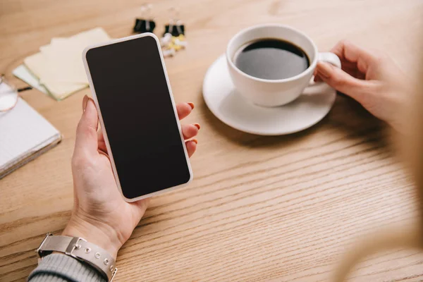 Cropped view of businesswoman holding cup of coffee while using smartphone at workplace — Stock Photo