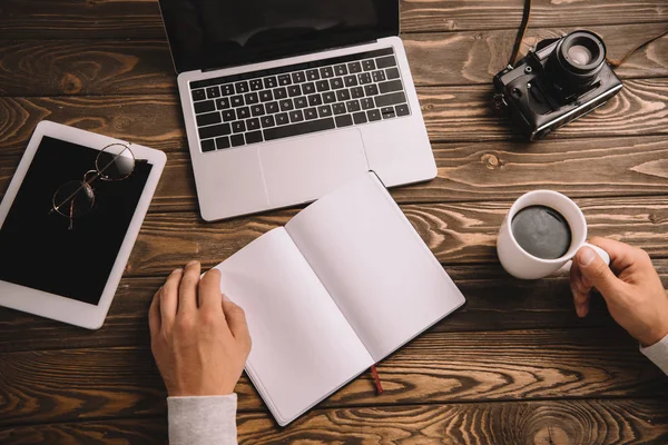 Vista ritagliata del blogger con notebook, tazza di caffè, dispositivi digitali e fotocamera retrò — Foto stock