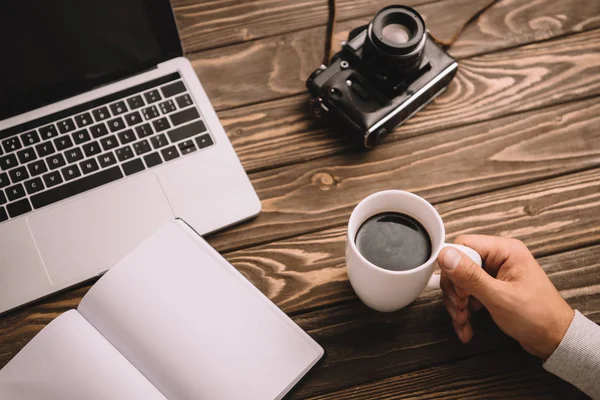 Cropped view of man with cup of coffee, notebook, laptop and retro photo camera — Stock Photo