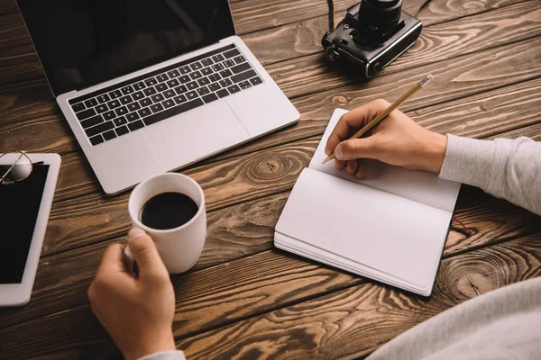 Vista recortada de la escritura del fotógrafo masculino en el cuaderno en la mesa con la taza de café, gadgets y cámara fotográfica retro - foto de stock