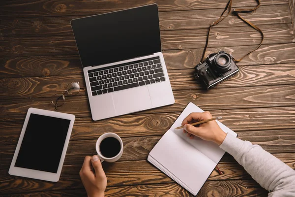 Vista recortada de la escritura freelancer masculino en el cuaderno en la mesa con taza de café, ordenador portátil, tableta y cámara de fotos retro - foto de stock