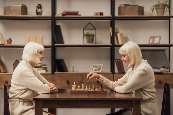 Sonrientes gemelas mayores jugando al ajedrez juntas en la sala de estar - foto de stock