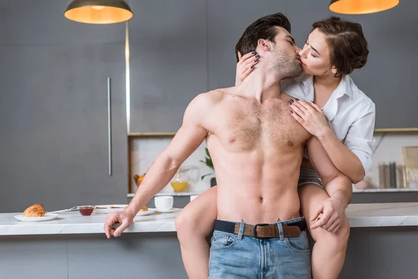 Adult couple kissing at kitchen while woman sitting on tabletop — Stock Photo