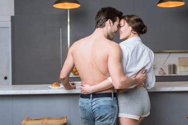 Visão traseira do namorado adulto e namorada abraçando, olhando nos olhos e sorrindo para a cozinha — Fotografia de Stock