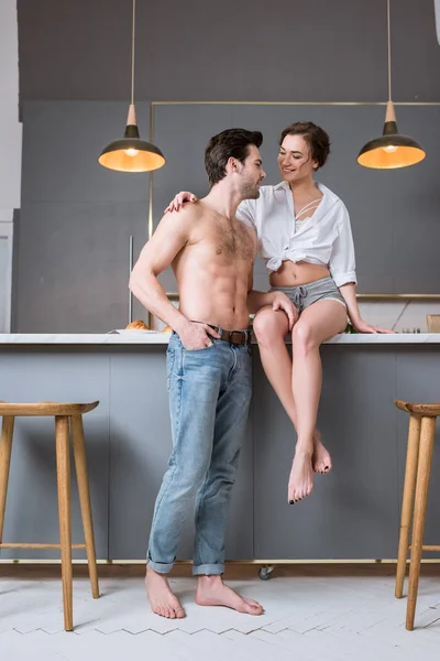 Bela mulher sentada na mesa e olhando bonito namorado sem camisa na cozinha — Fotografia de Stock