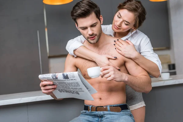 Schöne Freundin umarmt schönen hemdlosen Freund mit Tasse und Wirtschaftszeitung — Stockfoto