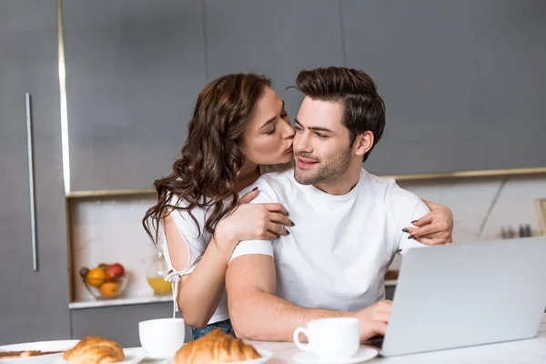 Mulher atraente beijando bochecha de namorado perto de laptop na cozinha — Fotografia de Stock