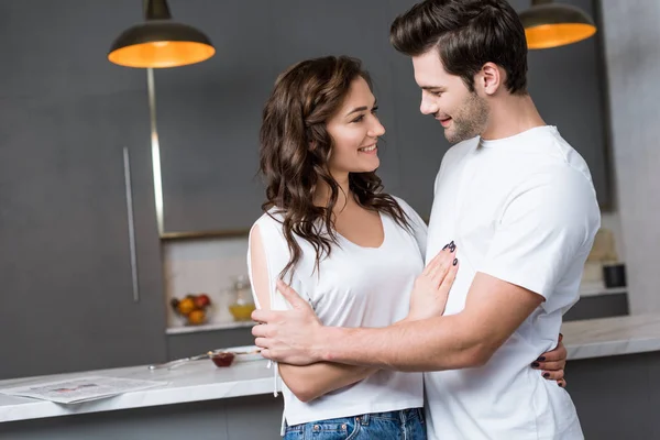 Mulher atraente abraçando e olhando para namorado bonito na cozinha — Fotografia de Stock