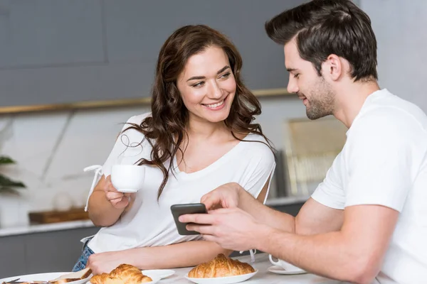 Petit ami gai tenant smartphone près de petite amie avec tasse de café — Photo de stock