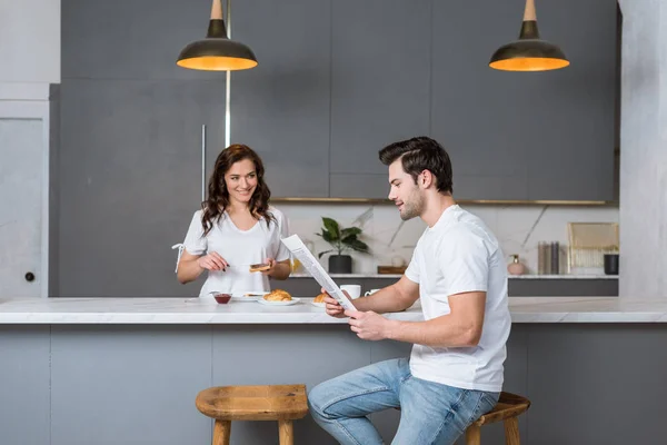 Beautiful girlfriend looking at handsome man reading newspaper in kitchen — Stock Photo