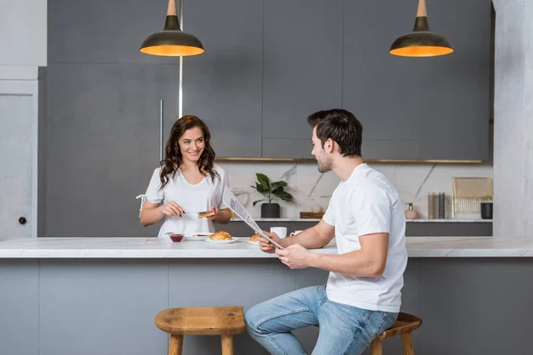 Beautiful girlfriend looking at handsome man holding newspaper in kitchen — Stock Photo