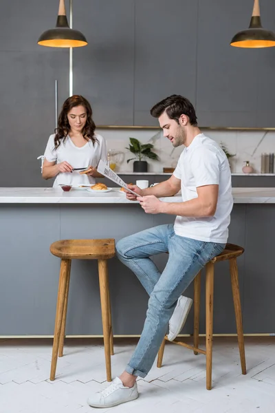 Homem bonito ler jornal perto de bela namorada na cozinha — Fotografia de Stock
