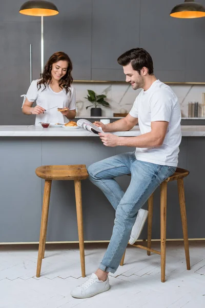 Homem bonito ler jornal perto de mulher atraente na cozinha — Fotografia de Stock