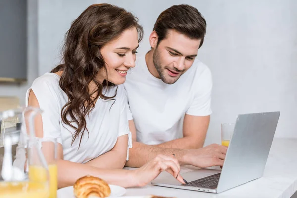 Alegre novio y novia sonriendo mientras usa el ordenador portátil en la cocina - foto de stock
