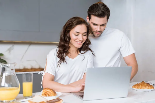 Mujer feliz mirando portátil cerca novio guapo en la cocina - foto de stock