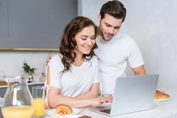 Feliz pareja usando el ordenador portátil cerca del desayuno sabroso en la cocina - foto de stock