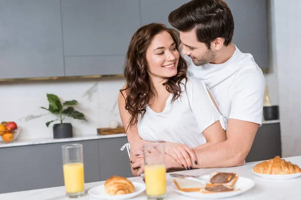 Casal feliz abraçando e sorrindo na cozinha perto de café da manhã saboroso — Fotografia de Stock