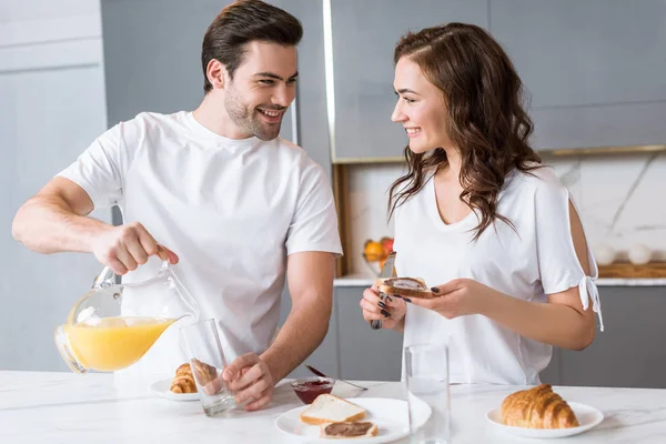 Bonito homem segurando jarro com suco de laranja e olhando para namorada na cozinha — Fotografia de Stock