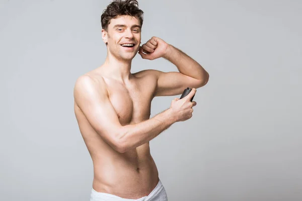 Веселый мужчина без рубашки, распыляющий дезодорант, изолированный на сером — стоковое фото