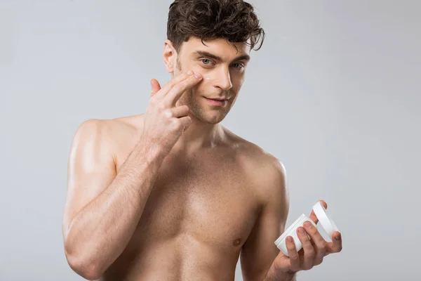 Muskulöser Mann, der Feuchtigkeitscreme auf das Gesicht aufträgt, isoliert auf grau — Stockfoto