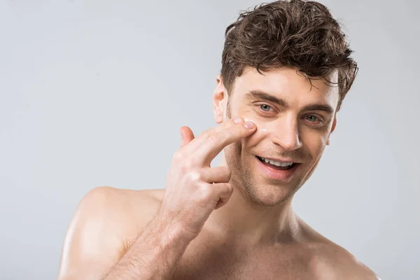 Lächelnder Mann, der kosmetische Creme auf das Gesicht aufträgt, isoliert auf grau — Stockfoto
