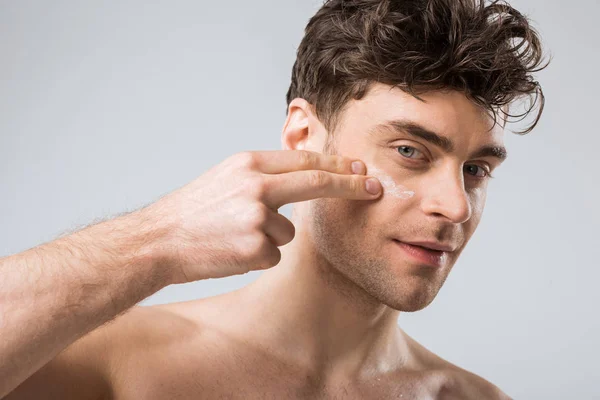 Giovane uomo che applica crema cosmetica sul viso, isolato su grigio — Foto stock