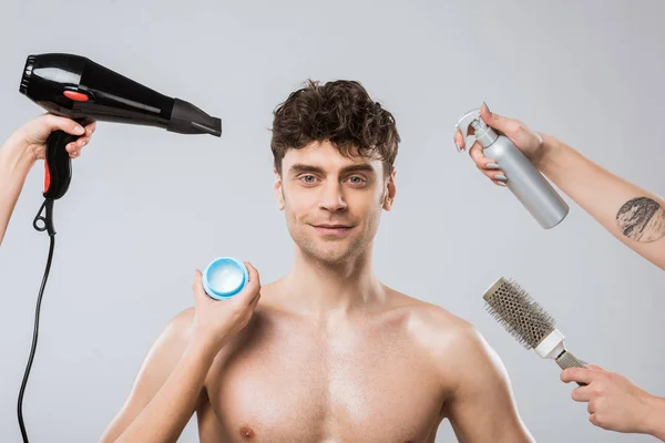 Hands of hairstylists holding hairdryer, spray, comb and styling gel naer smiling man, isolated on grey — Stock Photo