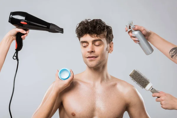 Mãos de cabeleireiros segurando secador de cabelo, spray, pente e estilo gel naer homem bonito, isolado em cinza — Fotografia de Stock