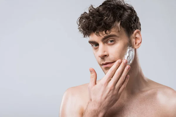 Bello giovane uomo che applica schiuma da barba sul viso, isolato su grigio — Foto stock