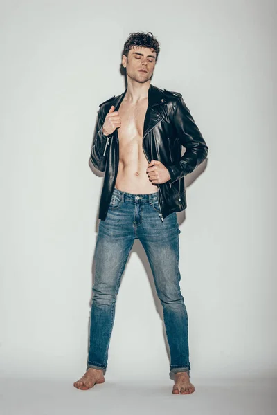 Sexy young man posing in jeans and black leather jacket on grey — Stock Photo