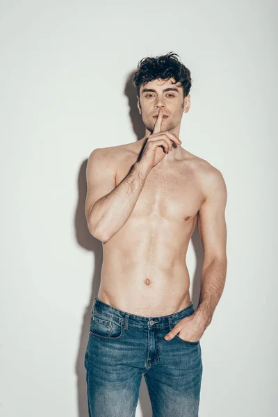 Sexy sin camisa hombre en jeans mostrando silencio símbolo mientras posando en gris - foto de stock