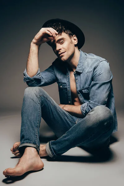 Модный сексуальный молодой человек позирует в джинсовой одежде и шляпе на темно-сером — стоковое фото