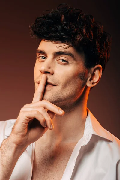 Apuesto hombre elegante en camisa blanca que muestra el símbolo del silencio, aislado en marrón - foto de stock