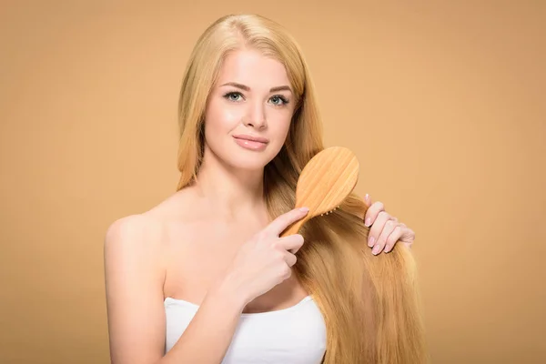 Menina loira atraente usando escova de cabelo e olhando para a câmera — Fotografia de Stock