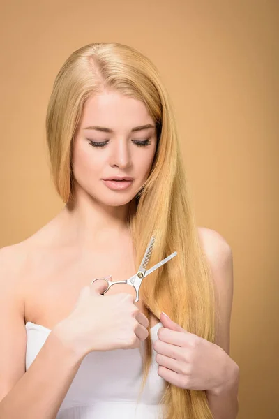 Attractive young woman cutting long blonde hair with scissors isolated on beige — Stock Photo