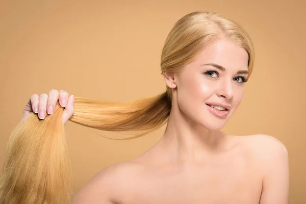 Bela mulher loira nua segurando cabelos longos e sorrindo para a câmera isolada no bege — Fotografia de Stock
